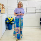Woman wearing a purple top and colorful patterned skirt standing in a room with a white wall and a blue flower arrangement.