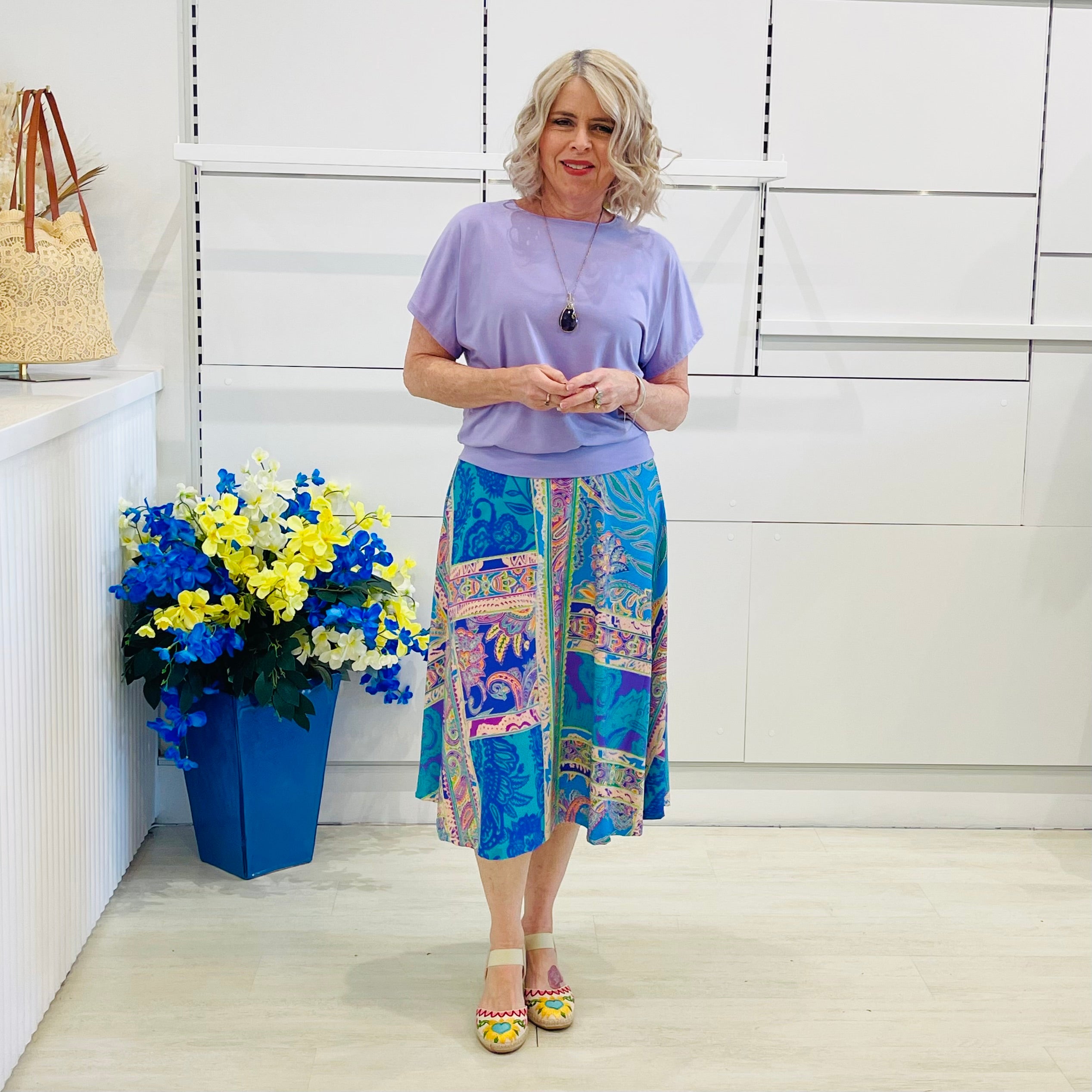 Woman wearing a purple top and colorful skirt standing next to a blue vase with flowers against a white wall.