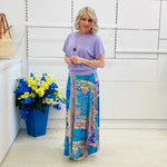 Woman wearing a purple top and colorful patterned skirt standing in front of a white wall with a blue flower pot.