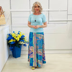 Woman wearing a light blue top and colorful patterned skirt standing next to a blue flowerpot with flowers.