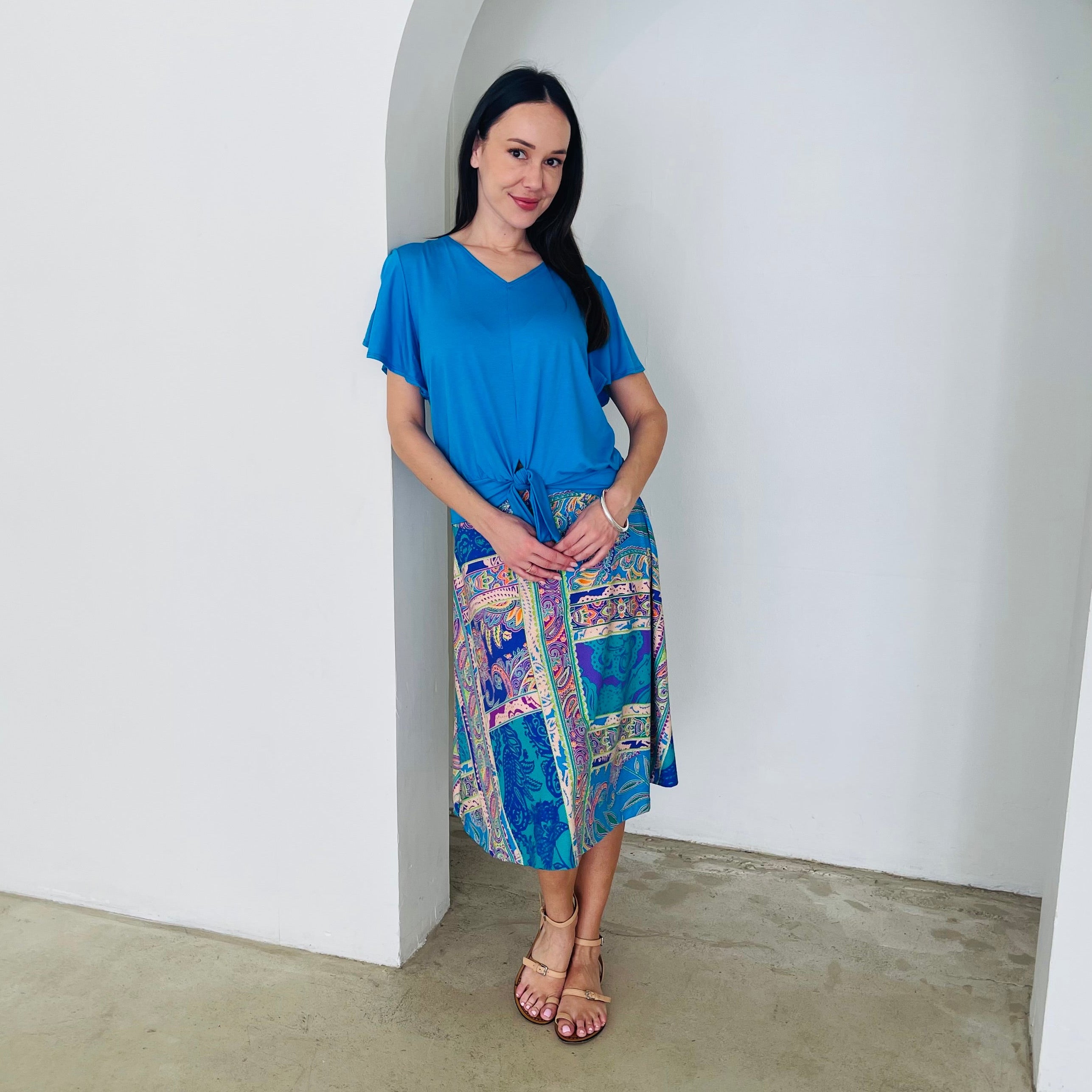 Woman wearing a blue top and colorful patchwork skirt standing against a white wall.