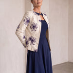 A woman models a cream-colored jacket with purple floral embroidery, worn over a navy blue midi dress in a minimalist, warm-toned indoor setting.