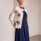 A woman models a cream-colored jacket with purple floral embroidery, worn over a navy blue midi dress in a minimalist, warm-toned indoor setting.