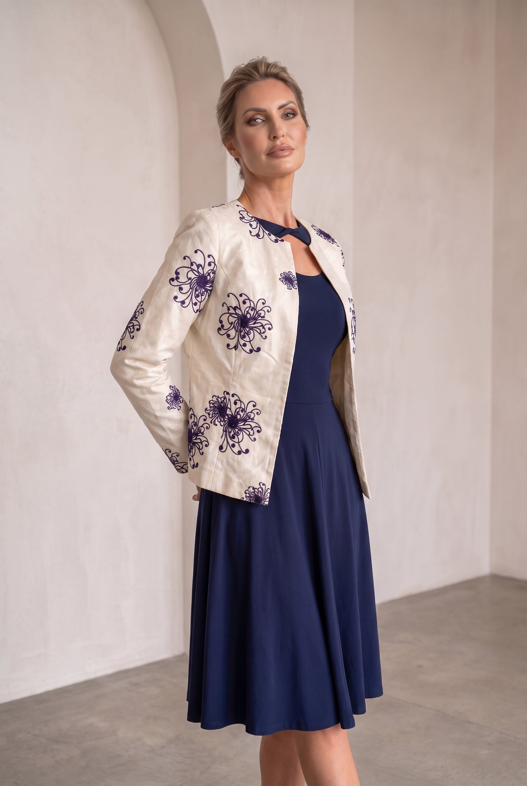 A woman models a cream-colored jacket with purple floral embroidery, worn over a navy blue midi dress in a minimalist, warm-toned indoor setting.