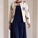 A woman models a navy blue midi dress with a keyhole neckline, styled with a cream-colored floral jacket and navy blue heels, set against a minimalist beige arched wall background.