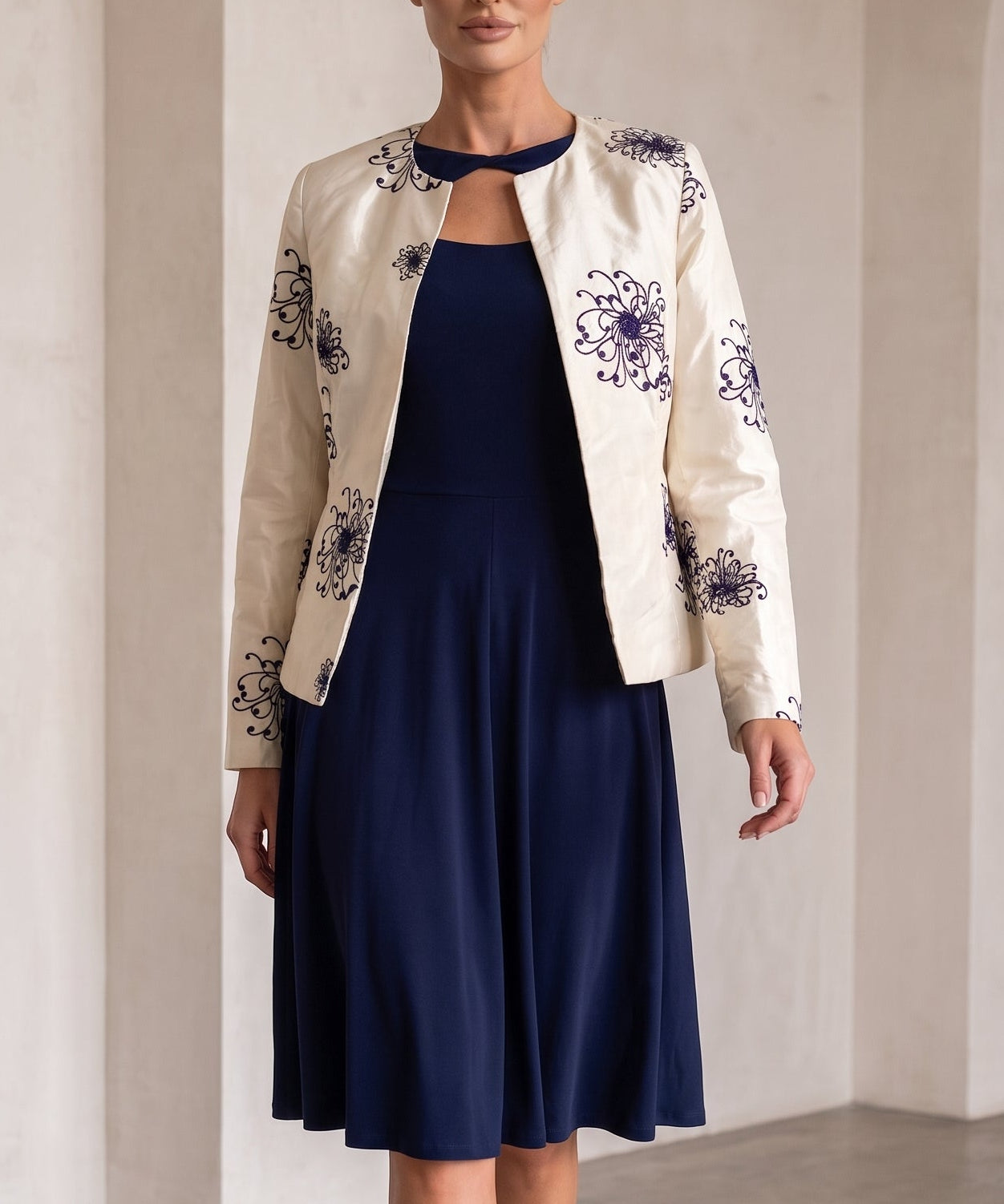 A woman models a navy blue midi dress with a keyhole neckline, styled with a cream-colored floral jacket and navy blue heels, set against a minimalist beige arched wall background.