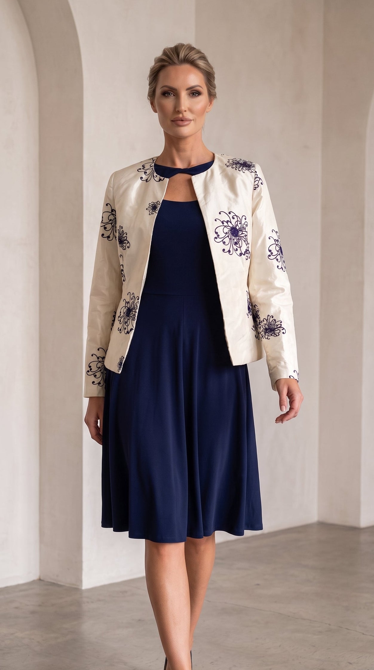 A woman models a navy blue midi dress with a keyhole neckline, styled with a cream-colored floral jacket and navy blue heels, set against a minimalist beige arched wall background.