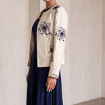 A woman models a cream-colored embroidered jacket over a navy blue dress in a minimalist, warm-toned indoor setting with arched walls.