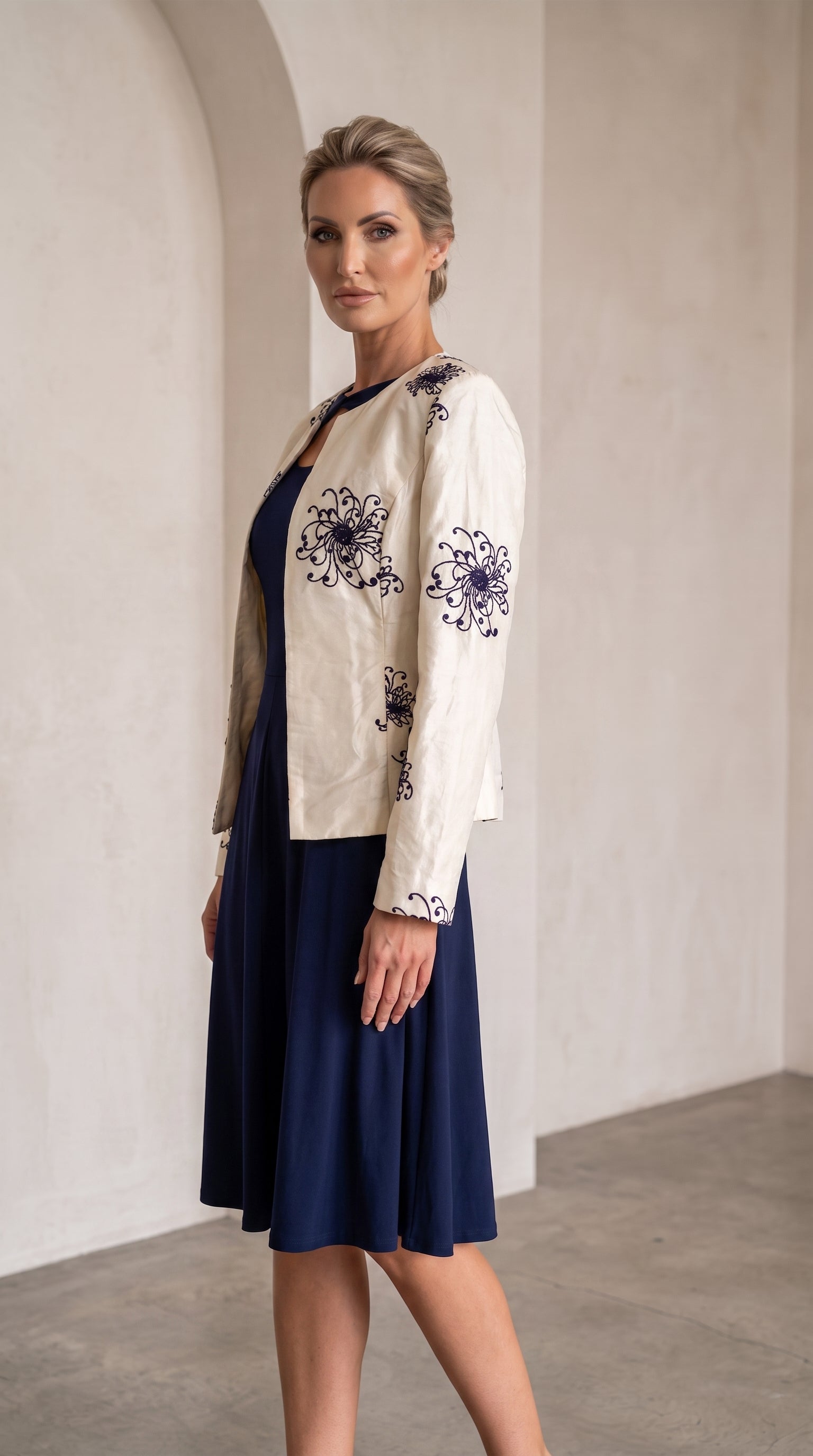 A woman models a cream-colored embroidered jacket over a navy blue dress in a minimalist, warm-toned indoor setting with arched walls.