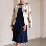 A woman models a navy blue midi dress paired with an off-white jacket featuring dark blue floral embroidery, standing in a minimalist, warm-toned interior with arched walls.