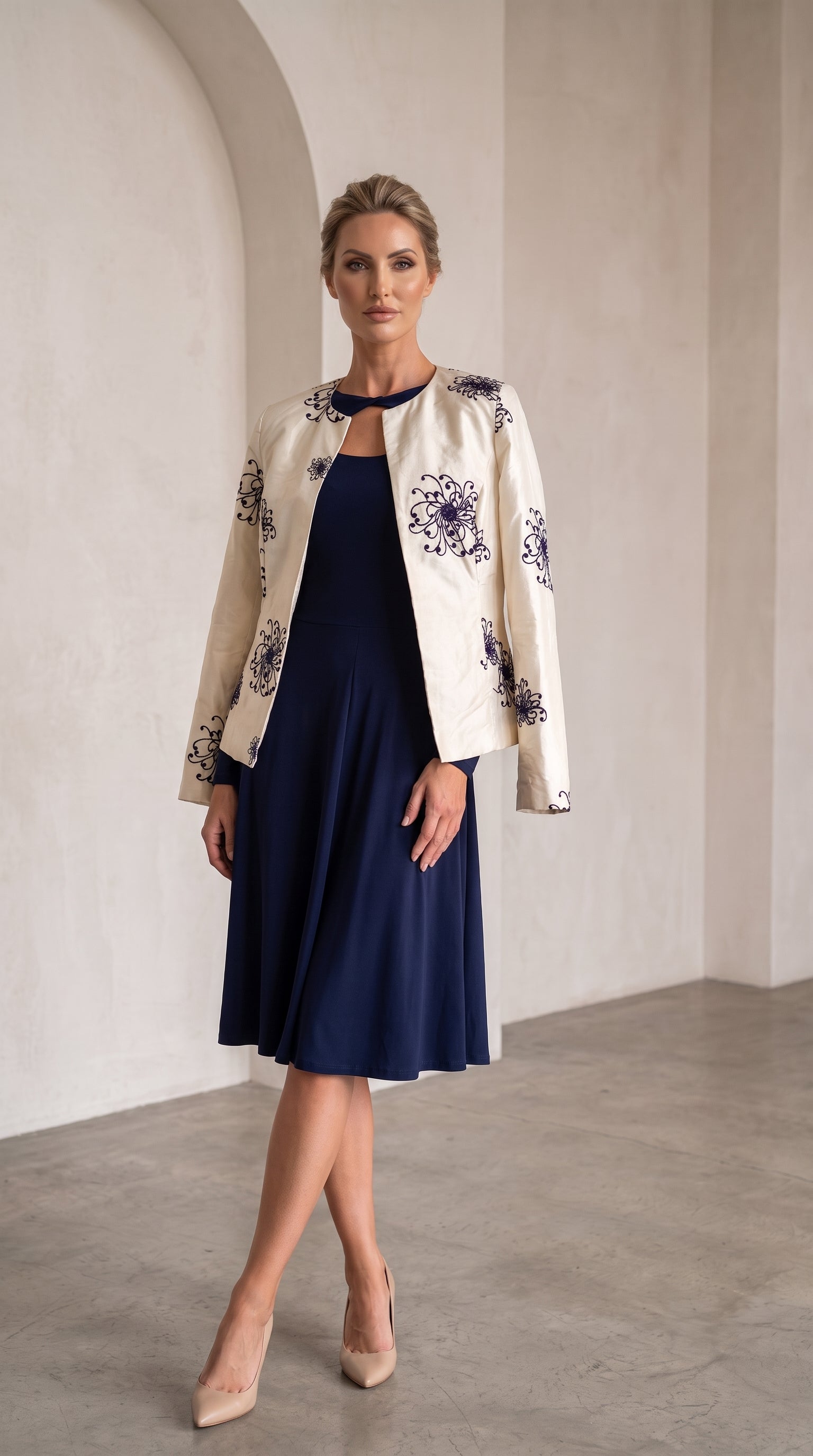 A woman models a navy blue midi dress paired with an off-white jacket featuring dark blue floral embroidery, standing in a minimalist, warm-toned interior with arched walls.