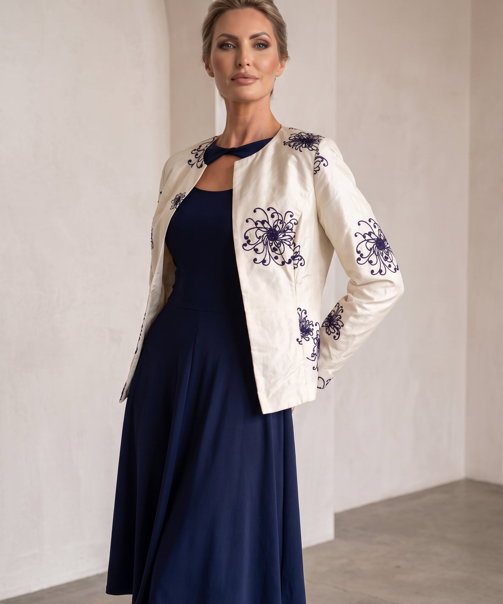 A woman models a navy blue midi dress paired with an off-white embroidered jacket. The jacket features dark blue floral embroidery on the sleeves and front.
