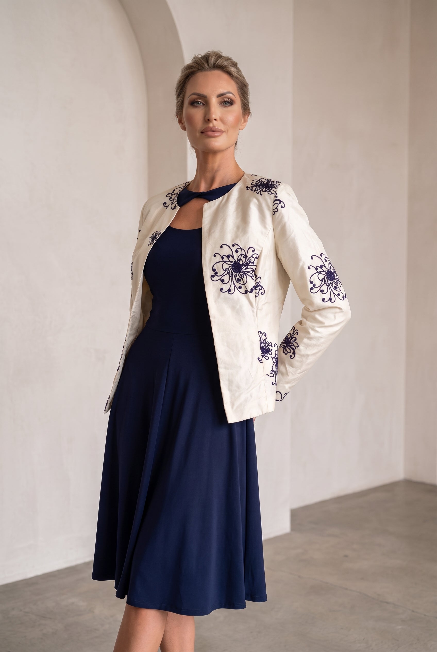 A woman models a navy blue midi dress paired with an off-white embroidered jacket. The jacket features dark blue floral embroidery on the sleeves and front.