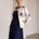 A woman models a navy blue midi dress paired with an off-white embroidered jacket. The jacket features dark blue floral embroidery on the sleeves and front.