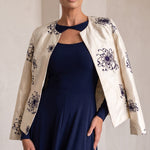 A woman modeling a navy blue maxi dress with a matching navy keyhole collar, styled with an open cream-colored jacket featuring dark blue floral embroidery, set against a neutral indoor background with arches.