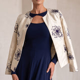 A woman modeling a navy blue maxi dress with a matching navy keyhole collar, styled with an open cream-colored jacket featuring dark blue floral embroidery, set against a neutral indoor background with arches.