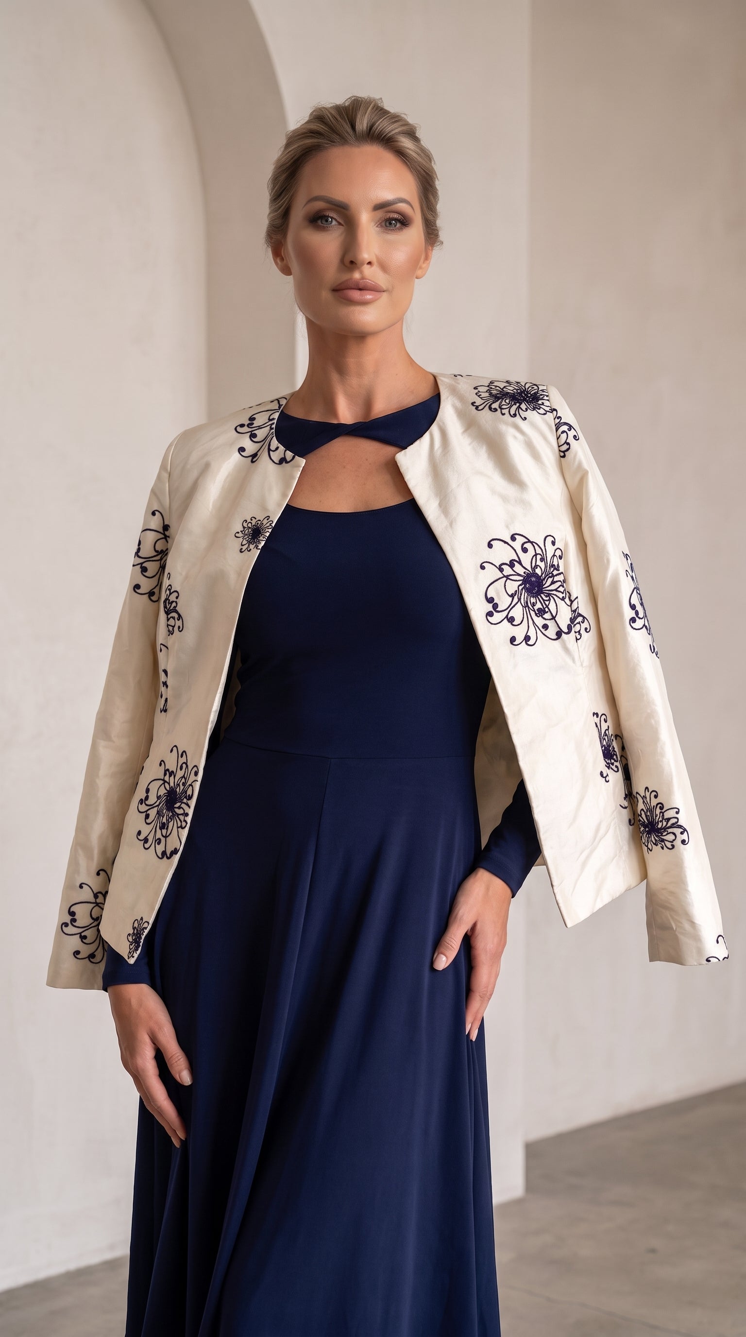 A woman modeling a navy blue maxi dress with a matching navy keyhole collar, styled with an open cream-colored jacket featuring dark blue floral embroidery, set against a neutral indoor background with arches.