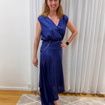 A woman wearing a sleeveless royal blue wrap-style maxi dress with a V-neckline, standing in an indoor room with white curtains, a patterned rug, and wooden flooring.