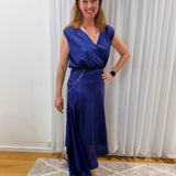 A woman wearing a sleeveless royal blue wrap-style maxi dress with a V-neckline, standing in an indoor room with white curtains, a patterned rug, and wooden flooring.