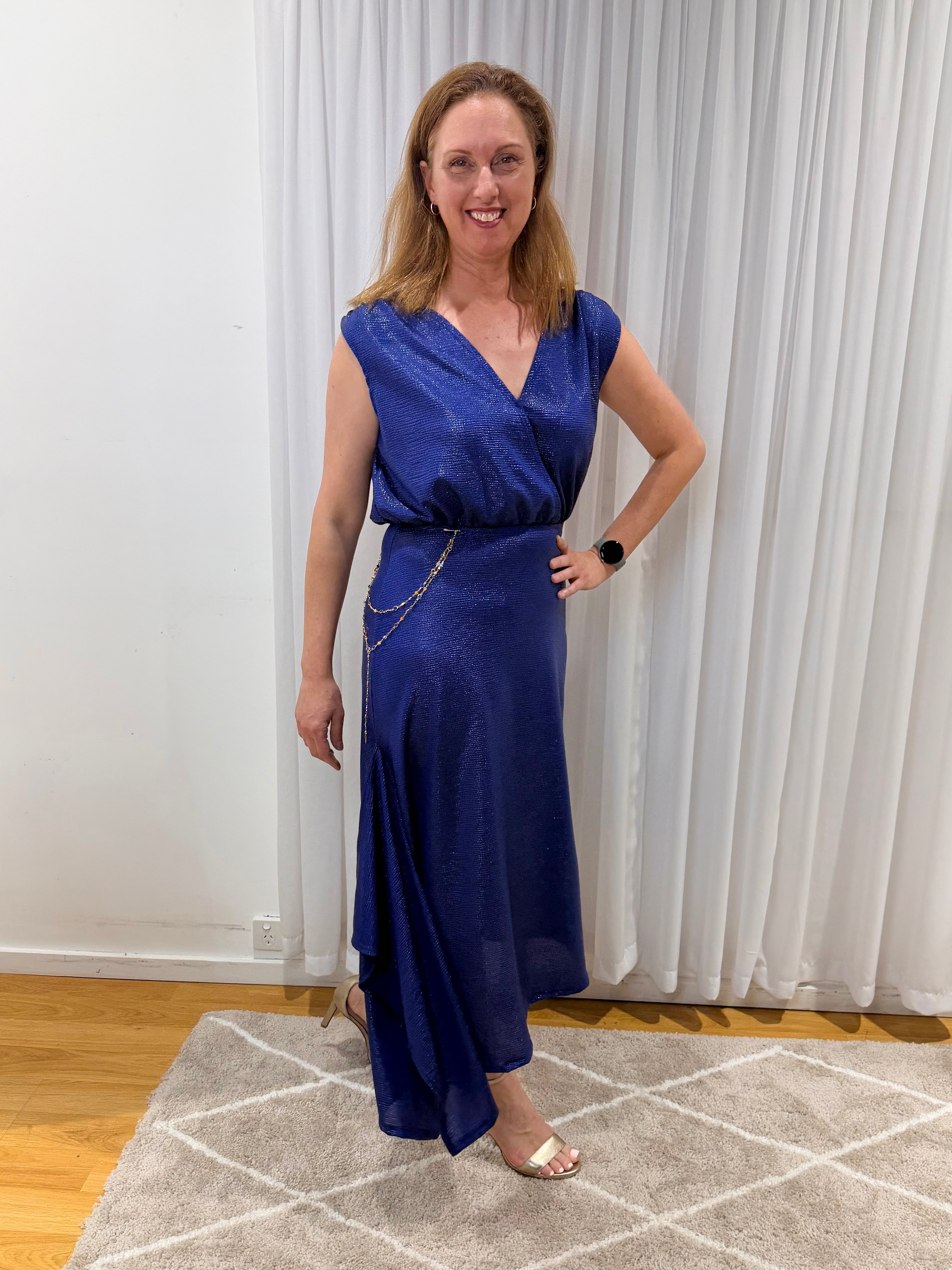 A woman wearing a sleeveless royal blue wrap-style maxi dress with a V-neckline, standing in an indoor room with white curtains, a patterned rug, and wooden flooring.