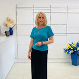Woman in a teal coloured top and black skirt standing in a room with white walls and decorative elements.