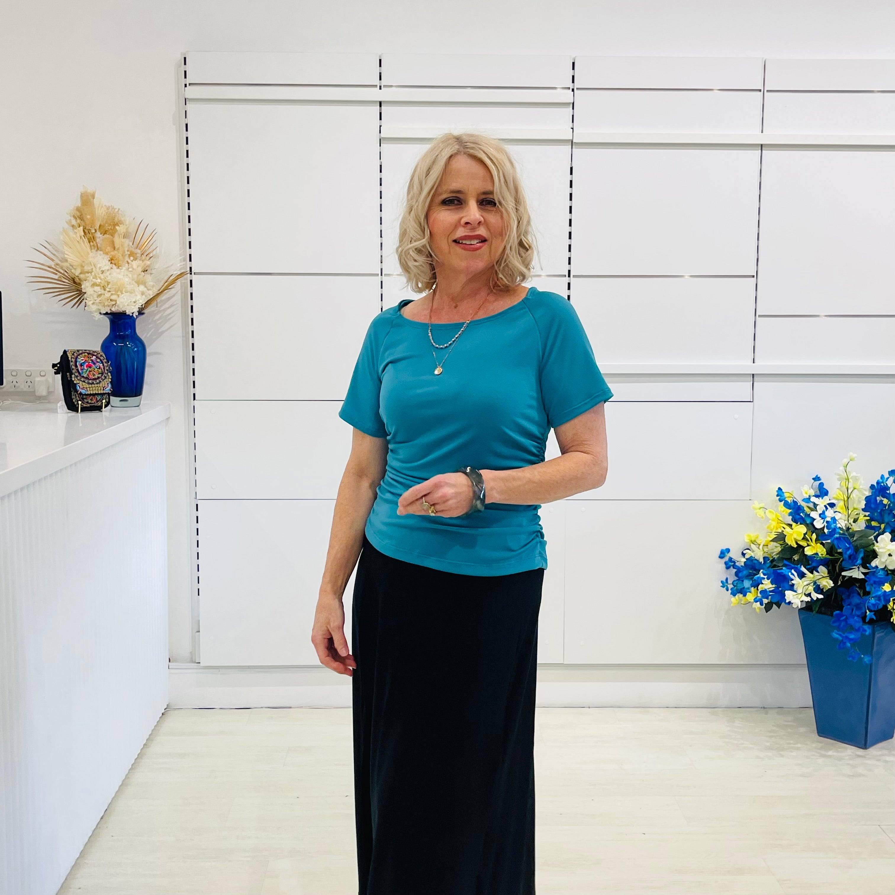 Woman in a teal coloured top and black skirt standing in a room with white walls and decorative elements.
