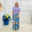 Woman in a light purple top and colorful patterned pants standing next to a blue flowerpot with flowers.