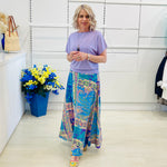 Woman wearing a purple top and colorful patterned skirt standing in a room with a white wall and a blue flower arrangement.