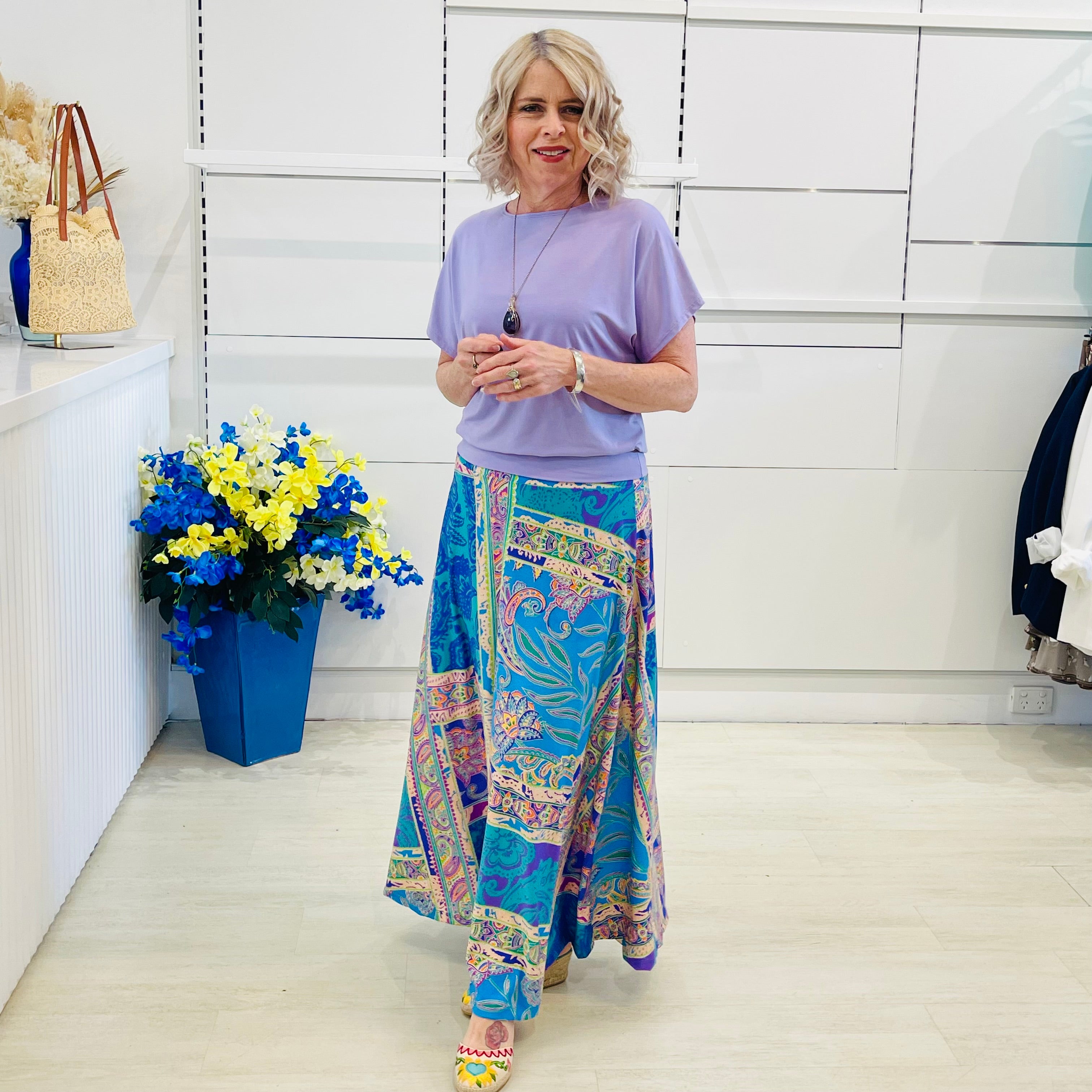 Woman wearing a purple top and colorful patterned skirt standing in a room with a white wall and a blue flower arrangement.