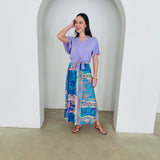 Woman wearing a purple top and colorful patterned skirt standing in a white archway.