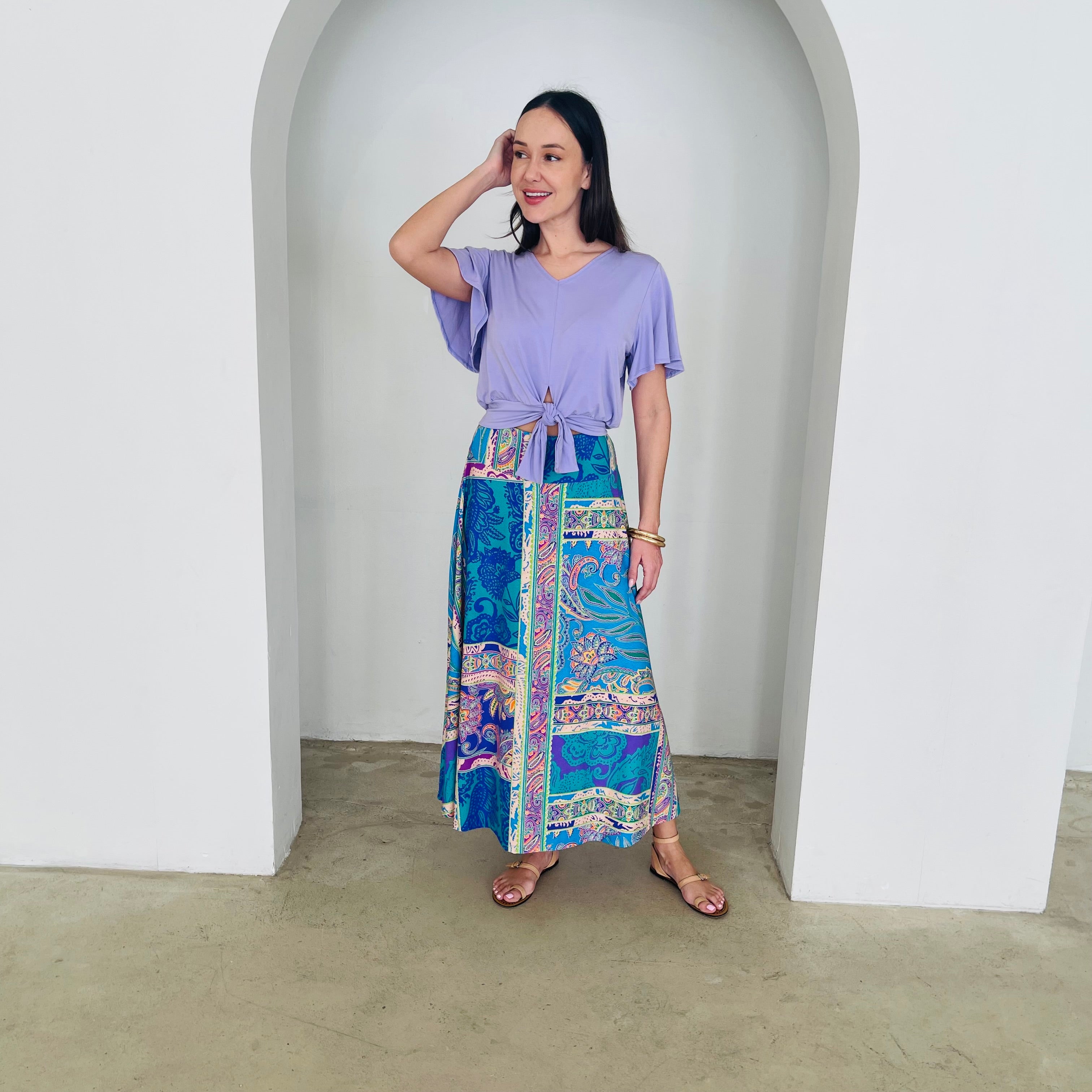 Woman wearing a purple top and colorful patterned skirt standing in a white archway.
