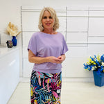 Woman wearing a purple top and colorful pants in a room with white walls and decorative elements.