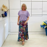 Woman wearing a light purple top and colorful skirt standing in a room with white walls and decorative elements.