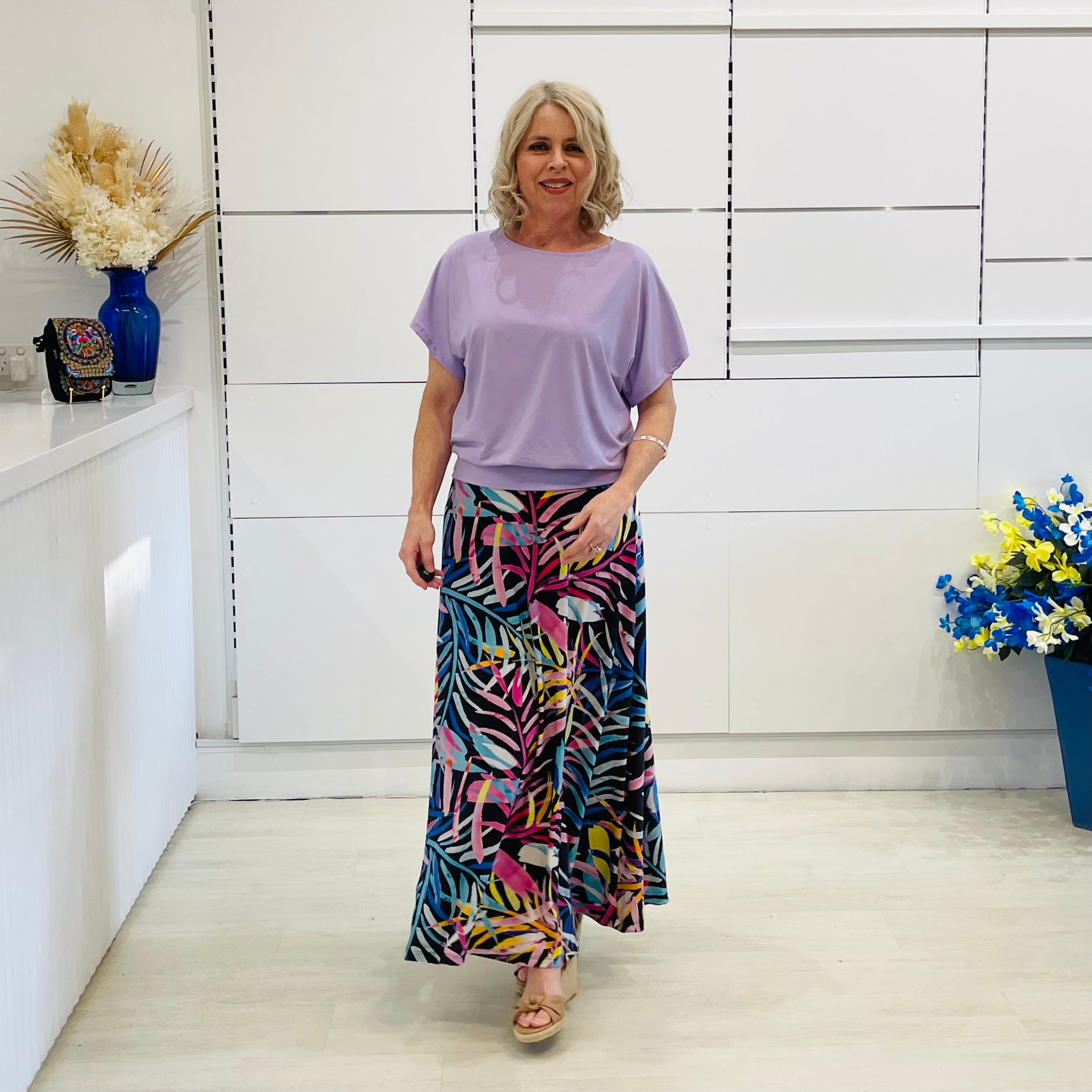 Woman wearing a light purple top and colorful skirt standing in a room with white walls and decorative elements.