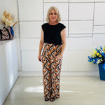 Woman wearing a black top and patterned pants standing in a room with white walls and a blue vase with flowers.