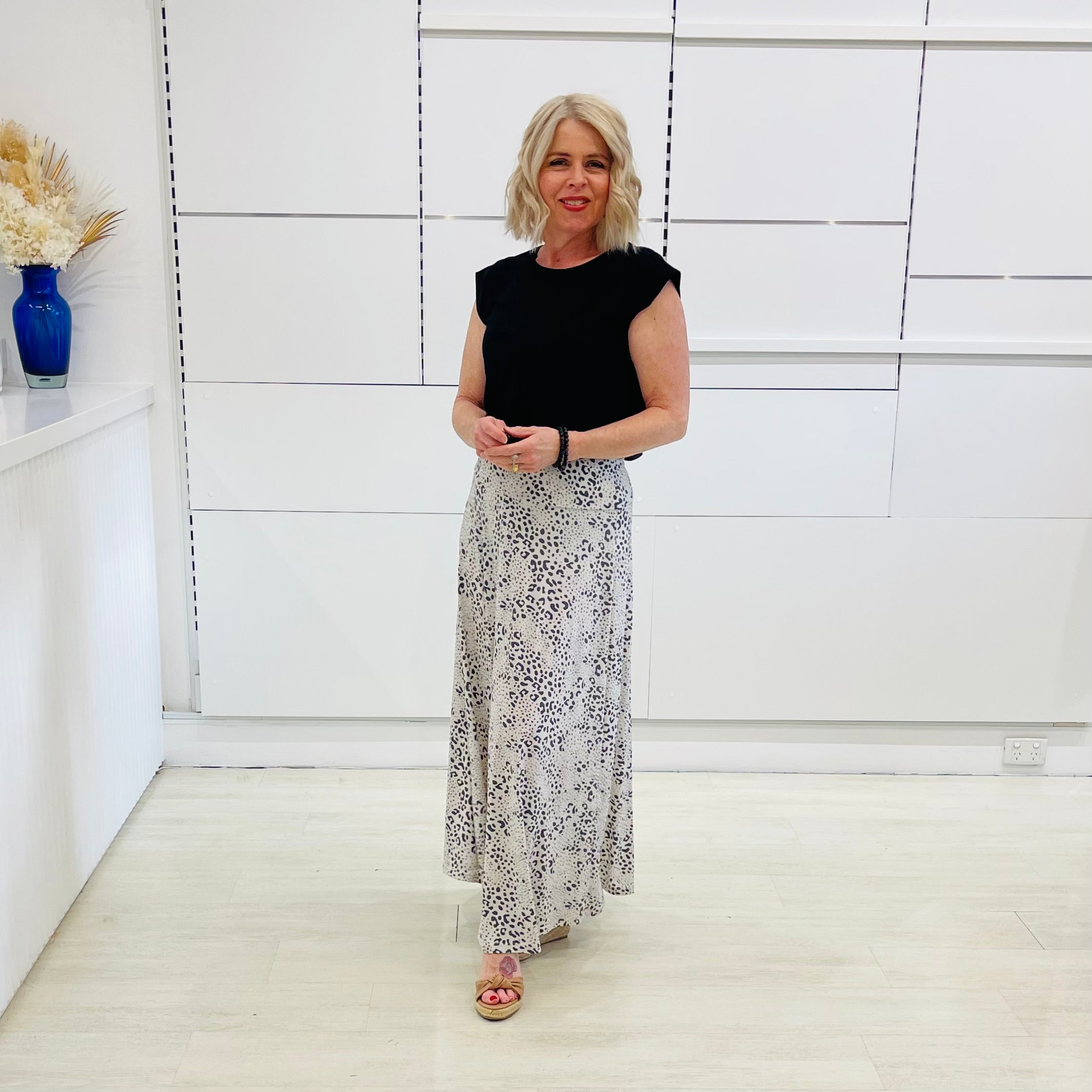 Woman in a black top and patterned skirt standing in a room with white walls and a vase on a shelf.