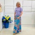 Woman wearing a purple top and colorful skirt standing next to a blue flowerpot with flowers.