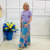 Woman wearing a purple top and colorful skirt standing next to a blue flowerpot with flowers.
