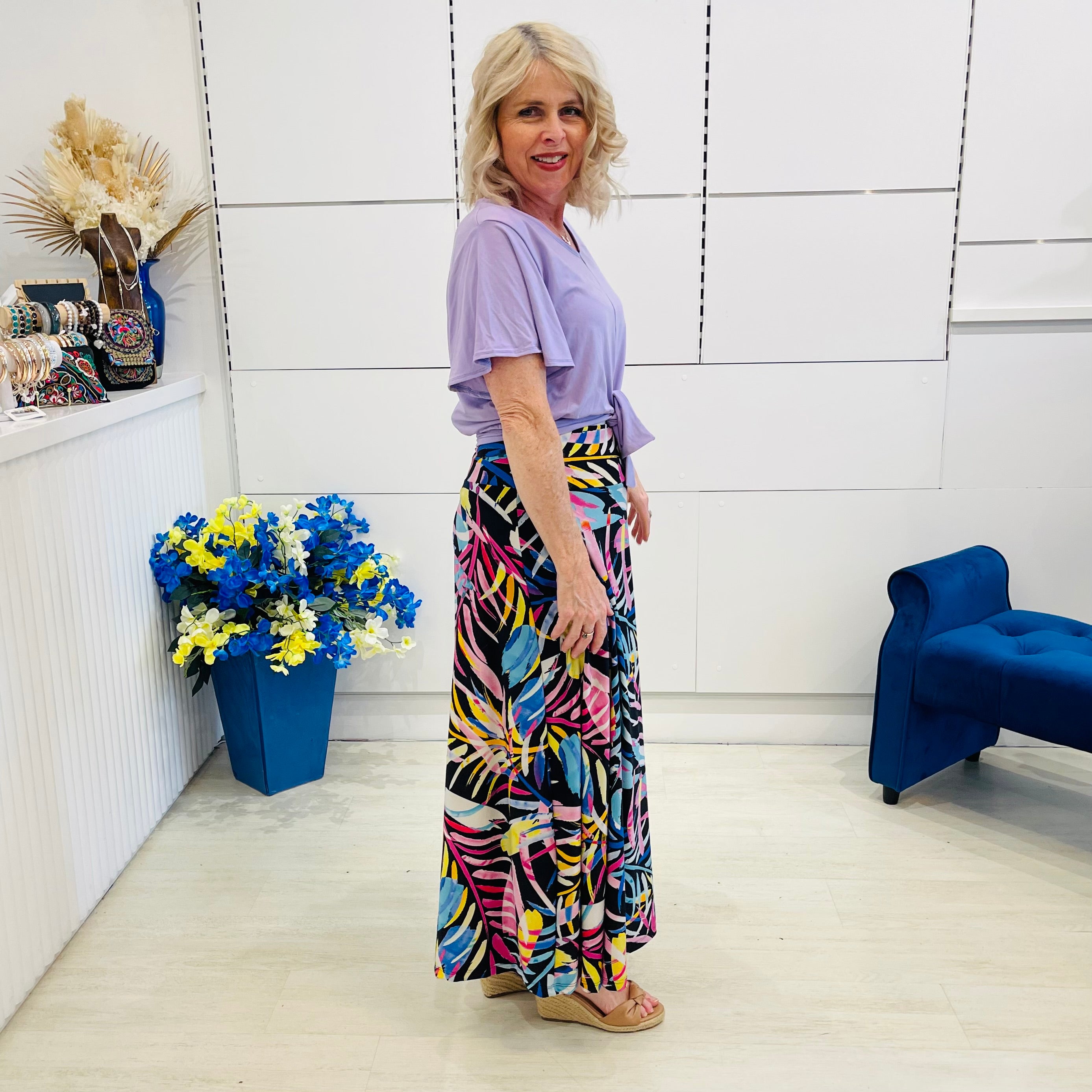 Woman in a colorful skirt and purple top standing in a room with a blue chair and flower arrangement.