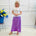 Woman wearing a white top and purple skirt standing in front of a white tiled wall with flowers in the background.