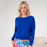 Woman wearing a blue top and colorful skirt in a room with a white wall and beige chair.