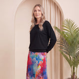 A woman stands in a studio setting wearing a black long-sleeve top and a colorful abstract-print midi skirt with blue, pink, red, and yellow tones. She is also wearing beige heeled ankle boots.