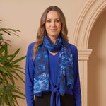 Woman wearing a blue top and black skirt with a blue patterned scarf, standing against a beige wall.