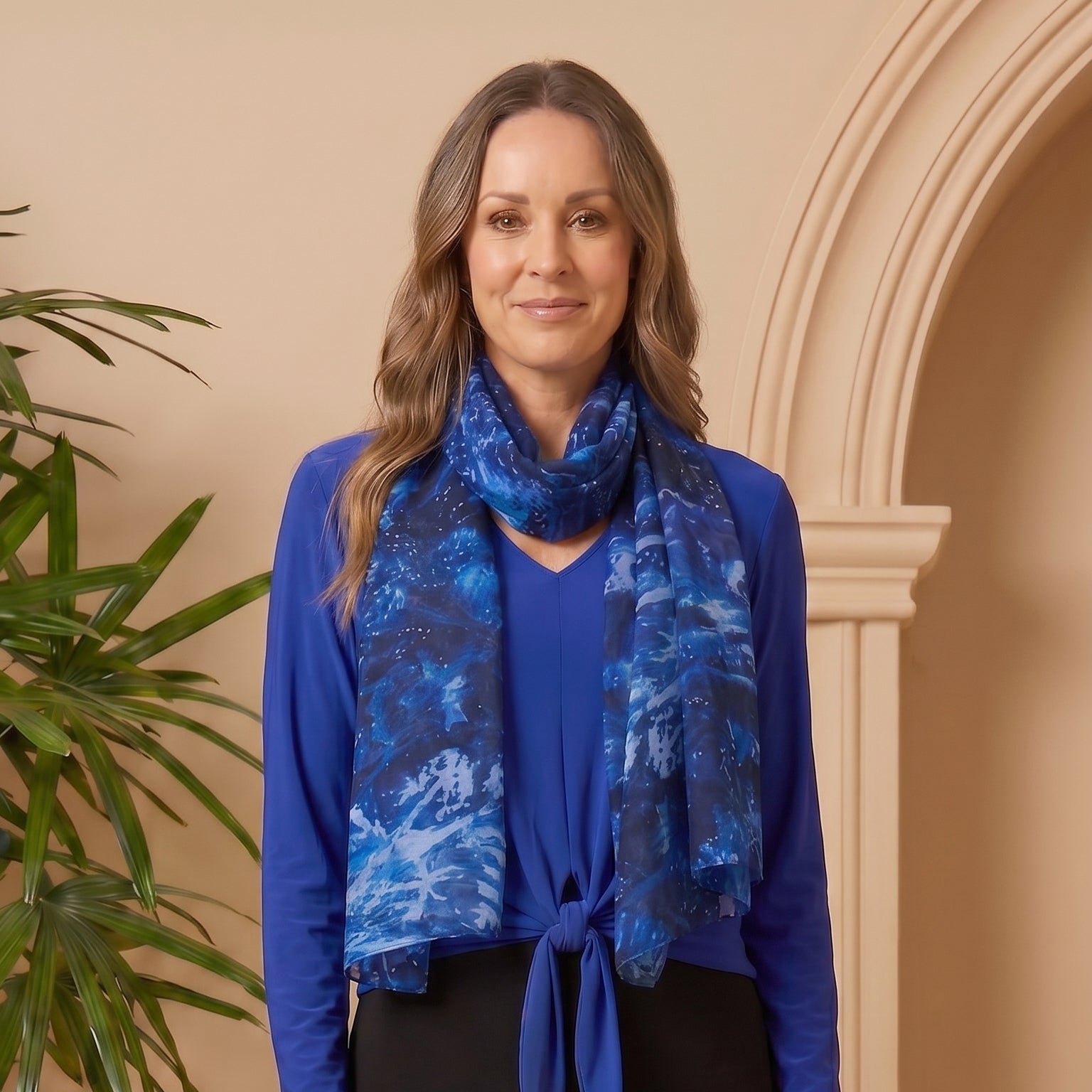 Woman wearing a blue top and black skirt with a blue patterned scarf, standing against a beige wall.