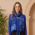 Woman wearing a blue top and black skirt with a blue patterned scarf, standing against a beige wall.