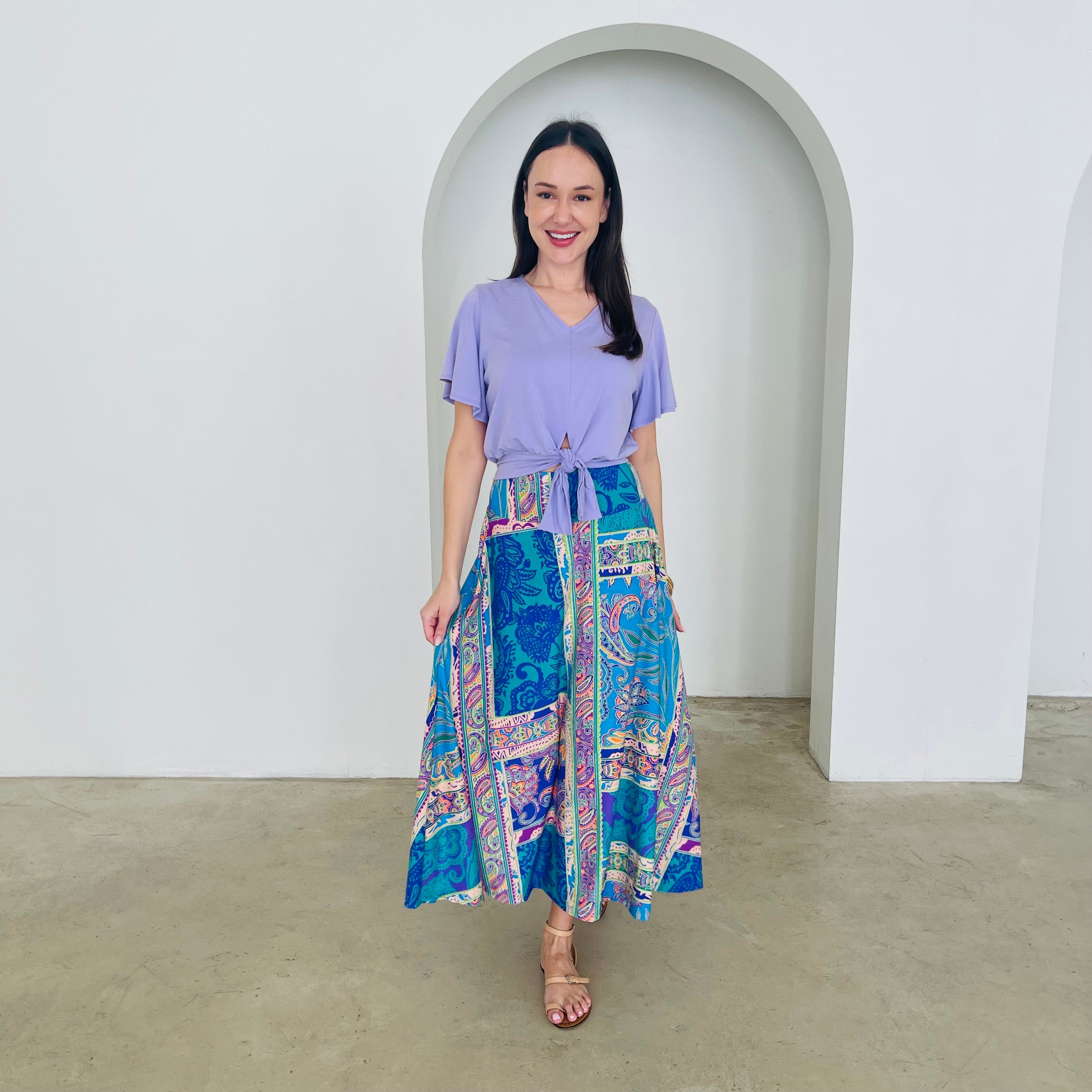 Woman wearing a purple top and colorful patterned skirt standing in front of a white archway.