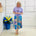 Woman wearing a purple top and colorful skirt standing next to a blue vase with flowers against a white wall.