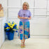 Woman wearing a purple top and colorful skirt standing next to a blue vase with flowers against a white wall.