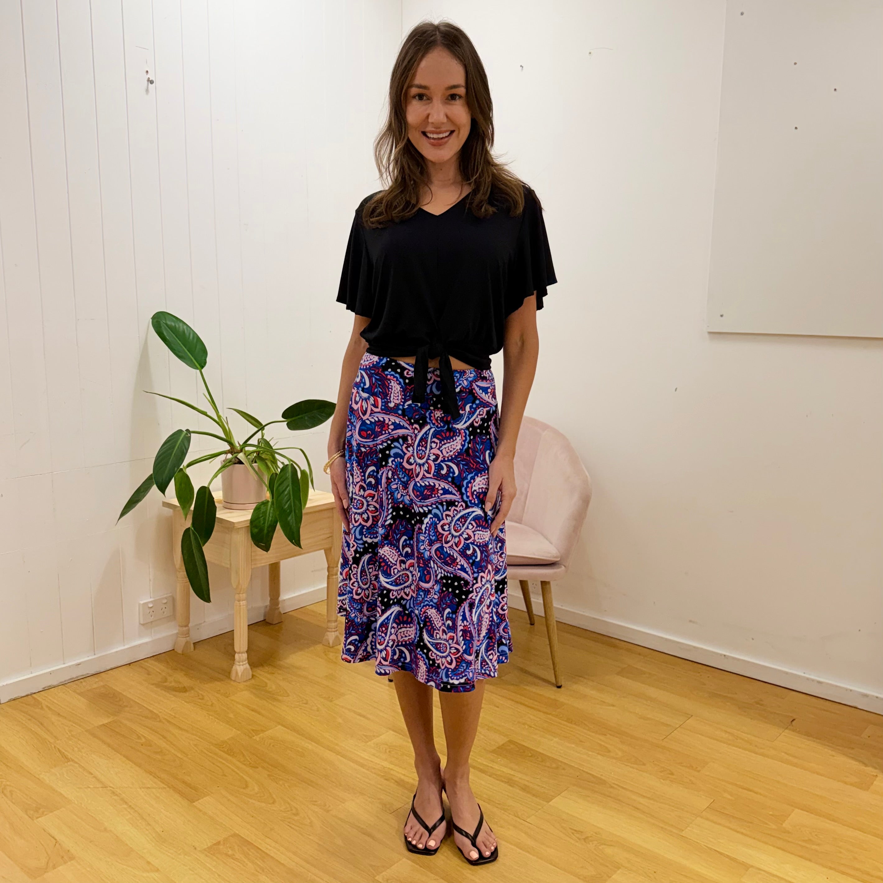 Woman wearing a black top and colorful patterned skirt standing in a room with a plant and chair.