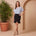 Woman in a light blue top and black skirt standing next to a potted plant against a beige wall.